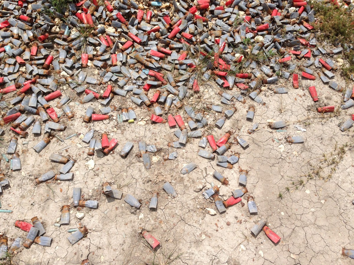 Color photograph of scattered red ammunition casings lying on a bare cracked ground