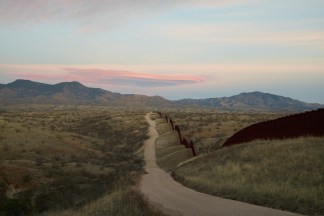 Color photograph of the US/Mexico border with a mountain range in the distance with pinkish blue skies