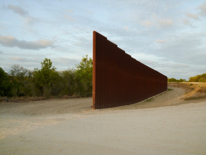 Color photograph of a tall metal fence segment ending in a bare dirt patch among low-lying trees and plants