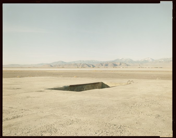Color photograph of a loading pit in the middle a desert with mountain range along the horizon with negative boarders around image
