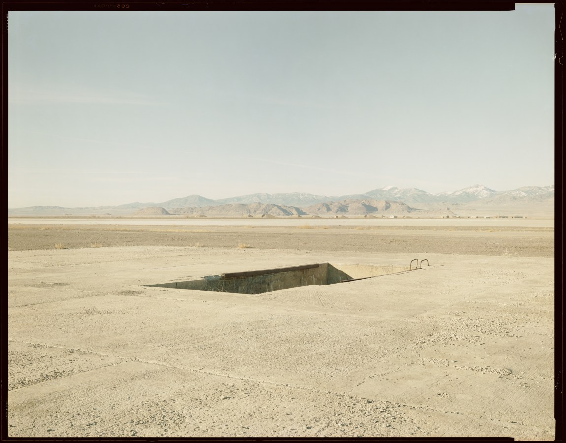 Color photograph of a loading pit in the middle a desert with mountain range along the horizon with negative boarders around image