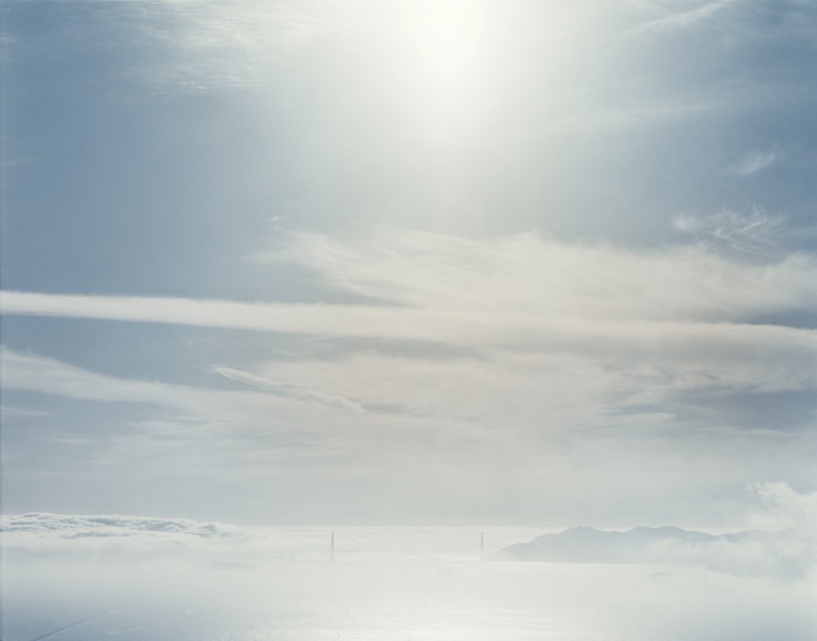 Color photograph of the distant Golden Gate Bridge through a low haze under a partly-clouded but still sunny sky