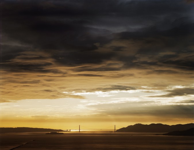 Color photograph of a bridge spanning a bay under a clearing of golden light through dark clouds