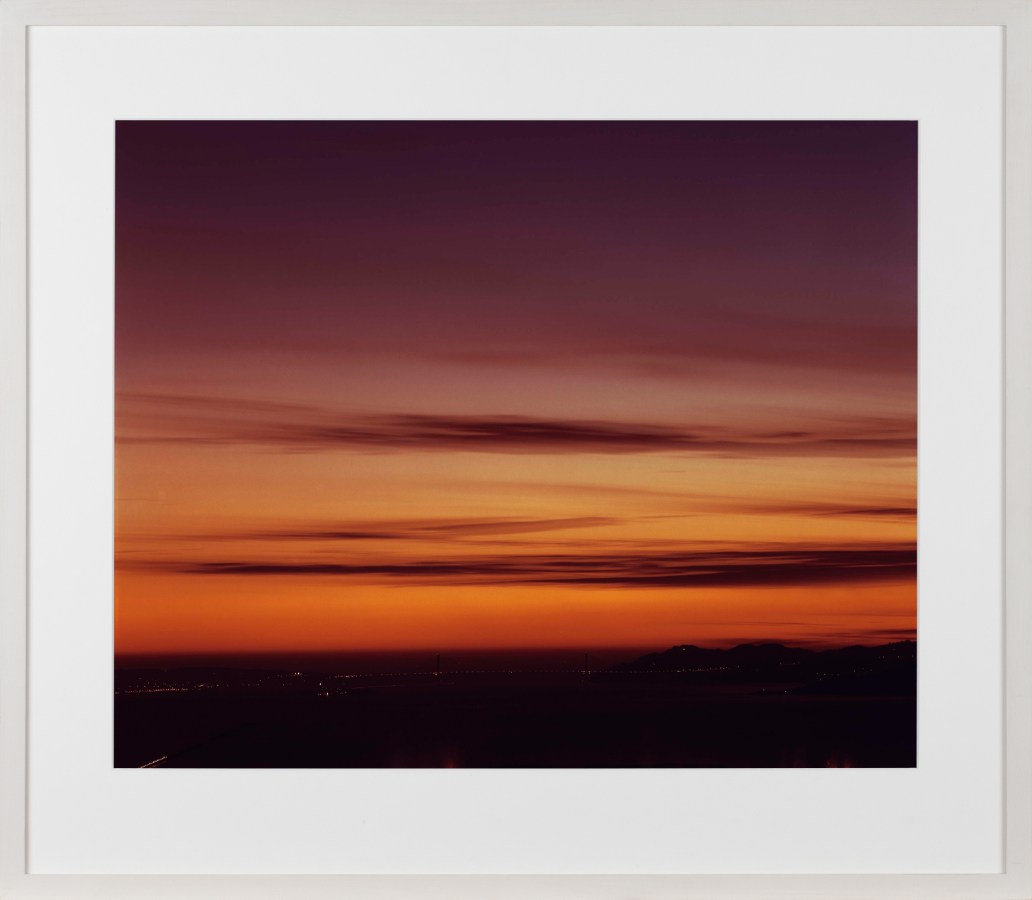 Color photograph of the Golden Gate Bridge and surrounding bay at sunset with a gradient sky of oranges and pinks framed in white