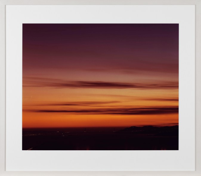 Color photograph of the Golden Gate Bridge and surrounding bay at sunset with a gradient sky of oranges and pinks framed in white