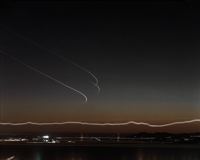 Long-exposure color photograph of the distant Golden Gate Bridge just after sunset under light trails of airplanes