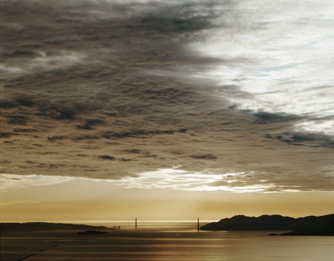 Color photograph of the distant Golden Gate Bridge at sunset under a cloudy sky