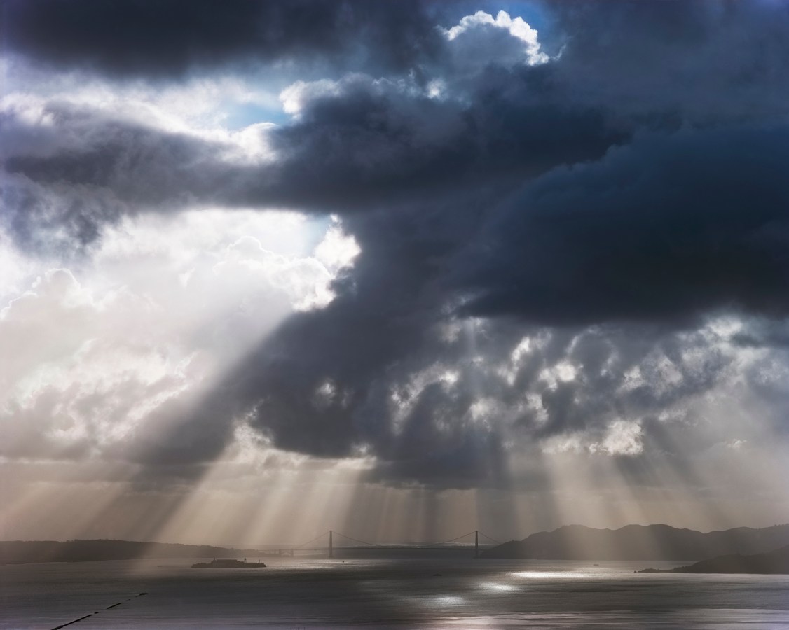 Color photograph of the distant Golden Gate Bridge under stark beams of sunlight coming through dark clouds