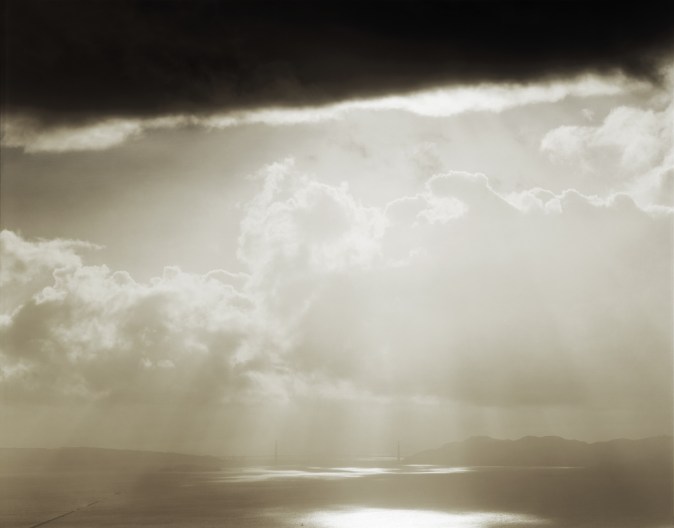 Color photograph of the distant Golden Gate Bridge under beams of sunlight breaking through a cloudy sky