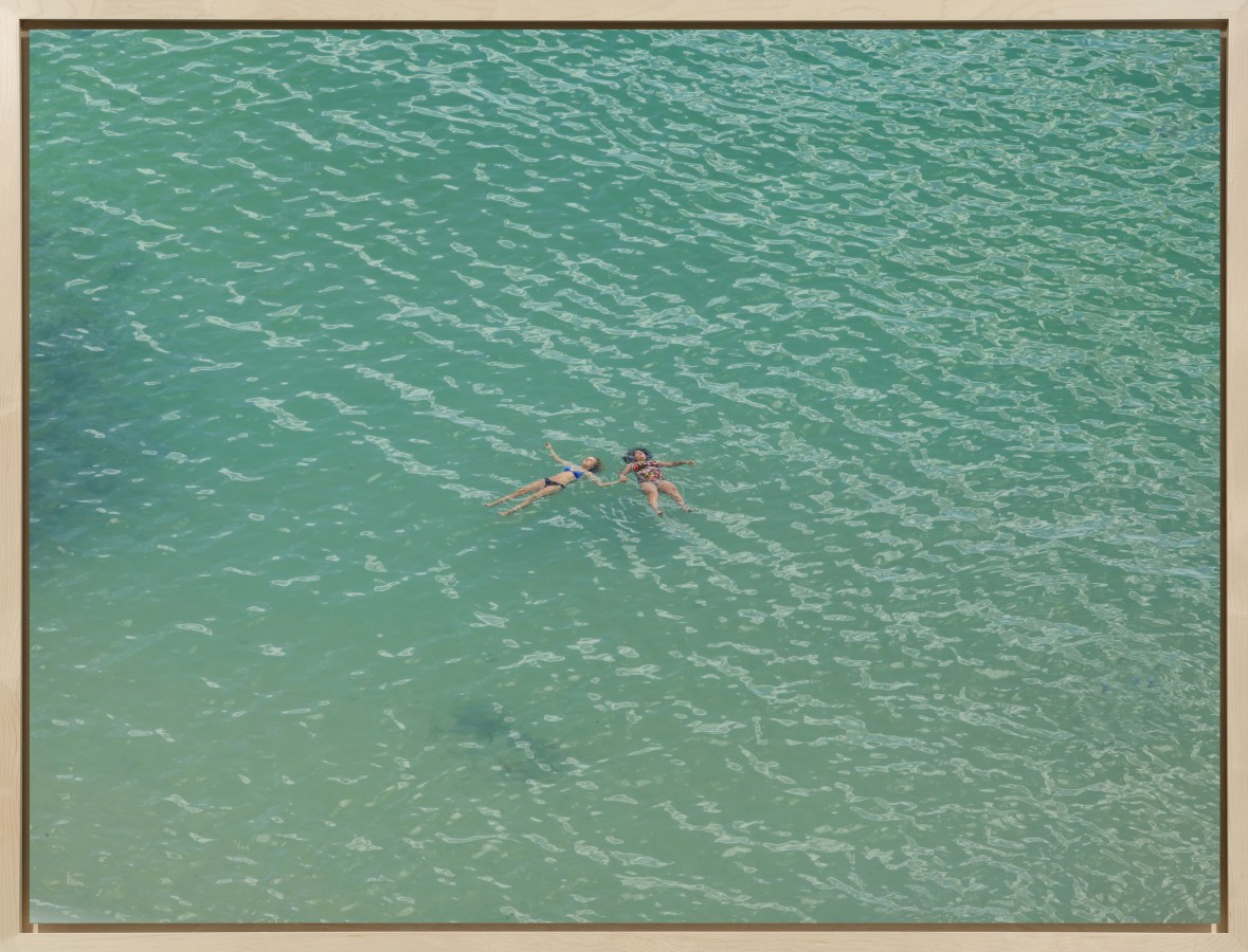 Color photograph of two women floating while holding hands on a calm, green ocean surface