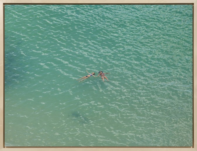 Color photograph of two women floating while holding hands on a calm, green ocean surface