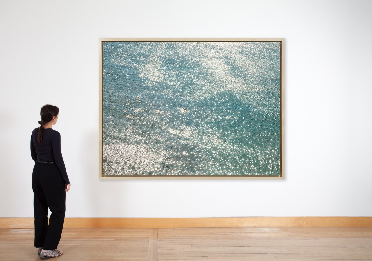 Color image of a figure viewing a color photograph depicting a single swimmer surrounded by water in a light wood frame on a white wall