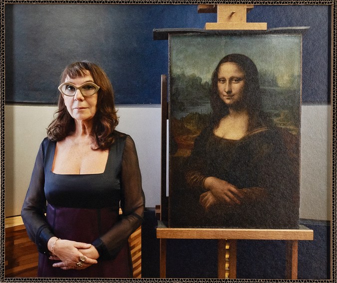 A color photograph depicts a woman in a black dress and glasses standing next to the Mona Lisa, Leonardo da Vinci’s famous painting of a woman.