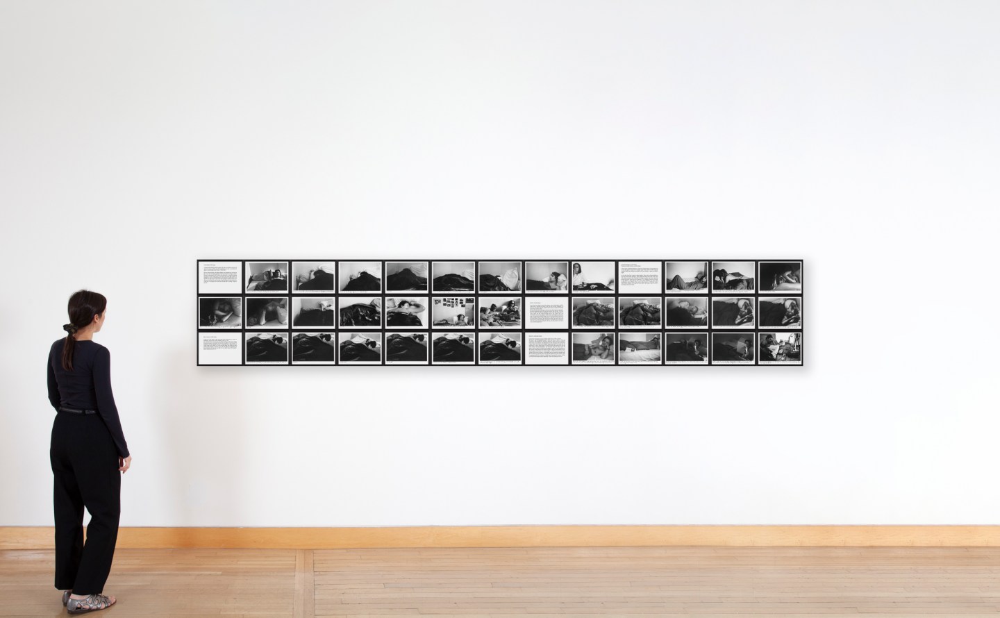 Color image of a figure viewing a suite of black and white photographs with text panels depicting and describing several subjects sleeping in a bed framed in black on a white wall