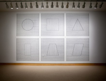 Color image of six gridded geometric squares with horizontal lines through each depicting a geometric form on white gallery wall