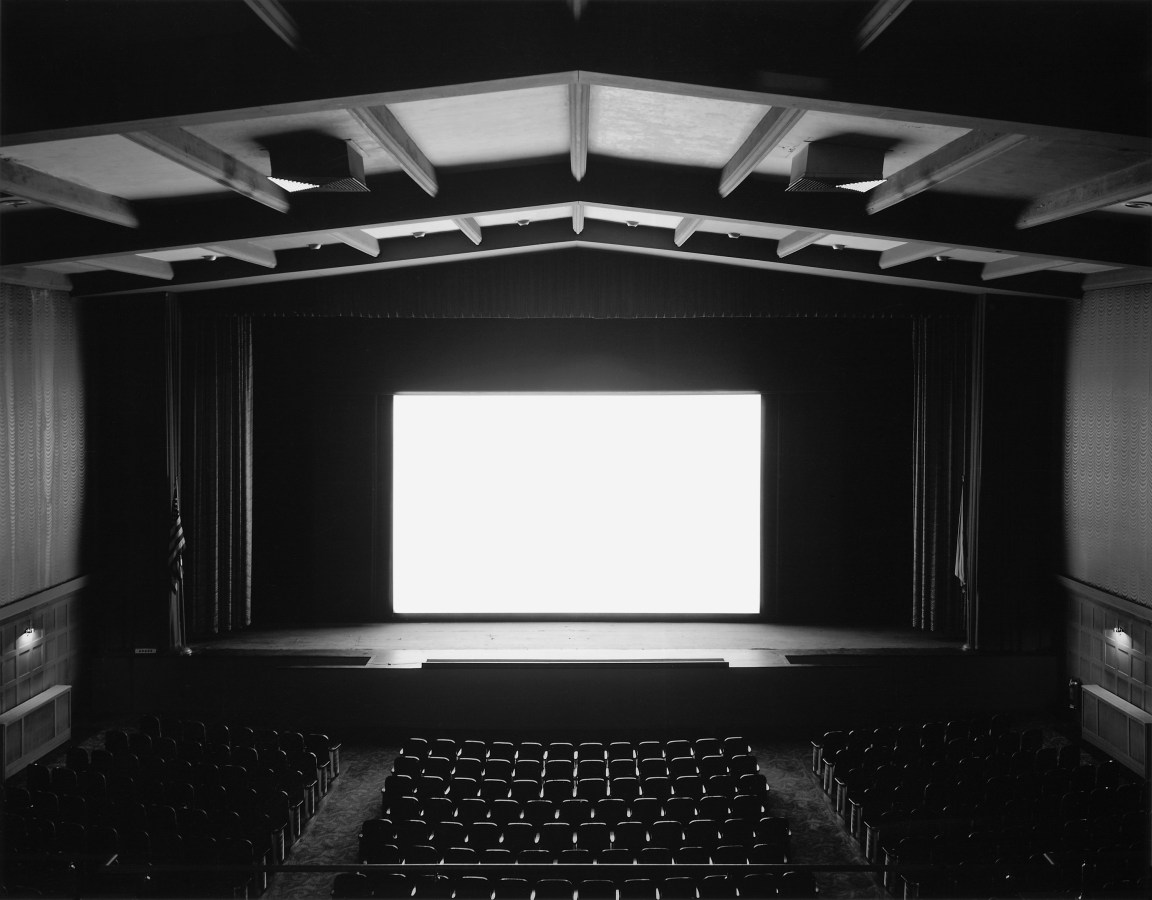 Black and white photograph of an empty theater with a blank white screen illuminating the space
