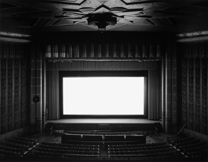 Black and white photograph of an empty theater with a blank white screen illuminating the space