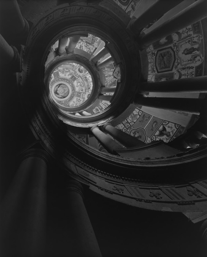 A black and white photograph of the view looking upward, at an elaborate spiral staircase.