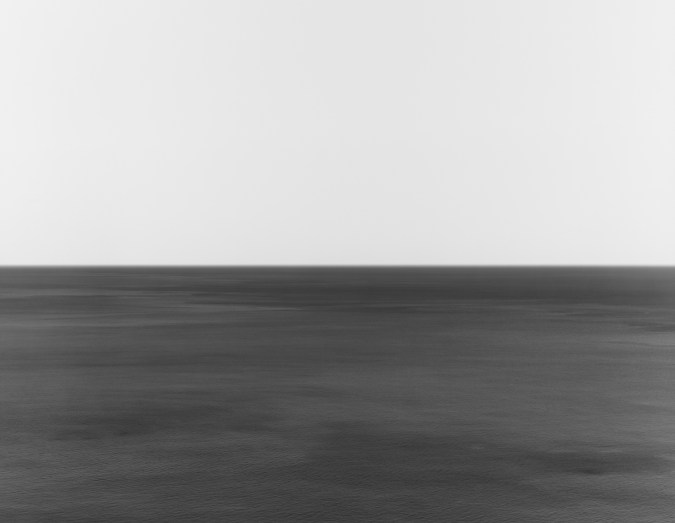 A black and white photograph of a seascape, with the horizon exactly in the middle of the frame. The water is rippled with some darker waves.