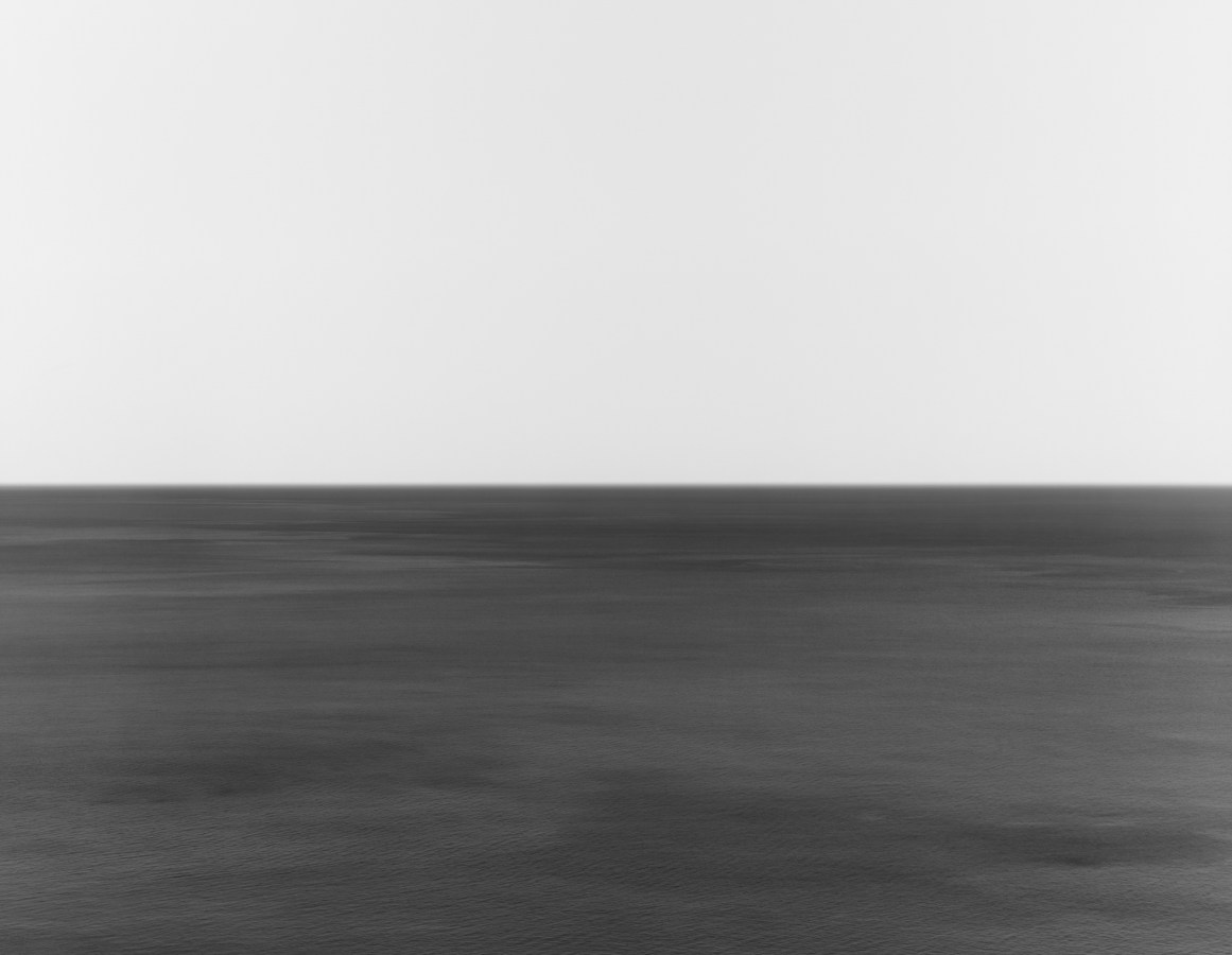 A black and white photograph of a seascape, with the horizon exactly in the middle of the frame. The water is rippled with some darker waves.