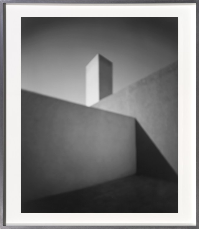 A framed black and white photograph of an outdoor courtyard, where two walls meet and a tower rises behind them.