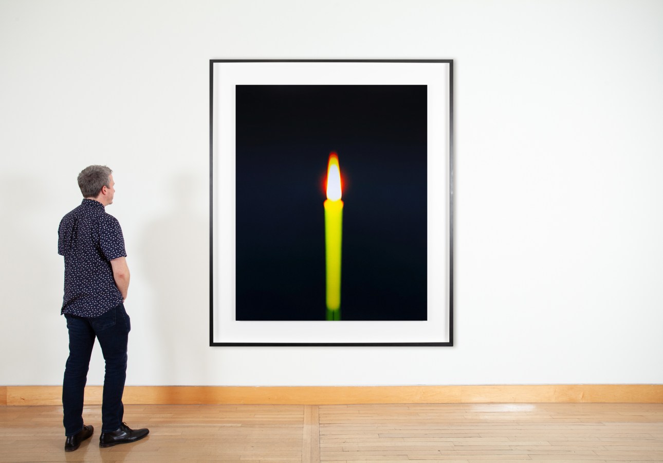 Color image of a figure viewing a color photograph depicting an out of focus burning candle framed in metal