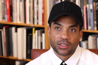 Video still depicting a figure wearing a black hat speaking in a library