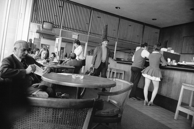 Black-and-white photograph of a man passing through a cafe area surrounded by tables and a counter