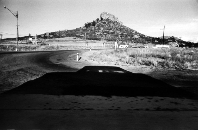 Black-and-white photograph of a roadside overlooking a rocky outcrop on a hill