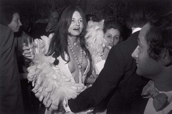 Black-and-white photograph of a woman in a low v-neck top with feathers and beaded necklaces holding a glass amidst a crowd of people